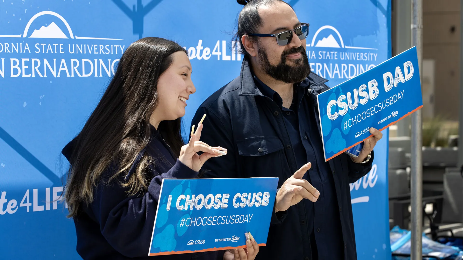 Two people holding up “Choose CSUSB Day” signs at a photo booth during the 2024 event