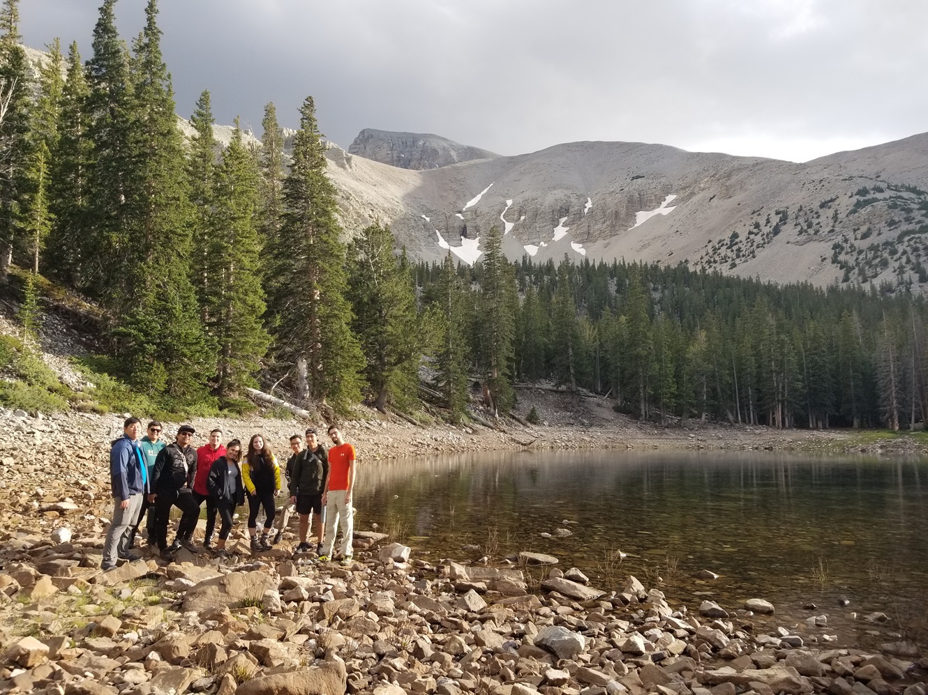 Group of 10 standing by lake with a mountain with snow behind.